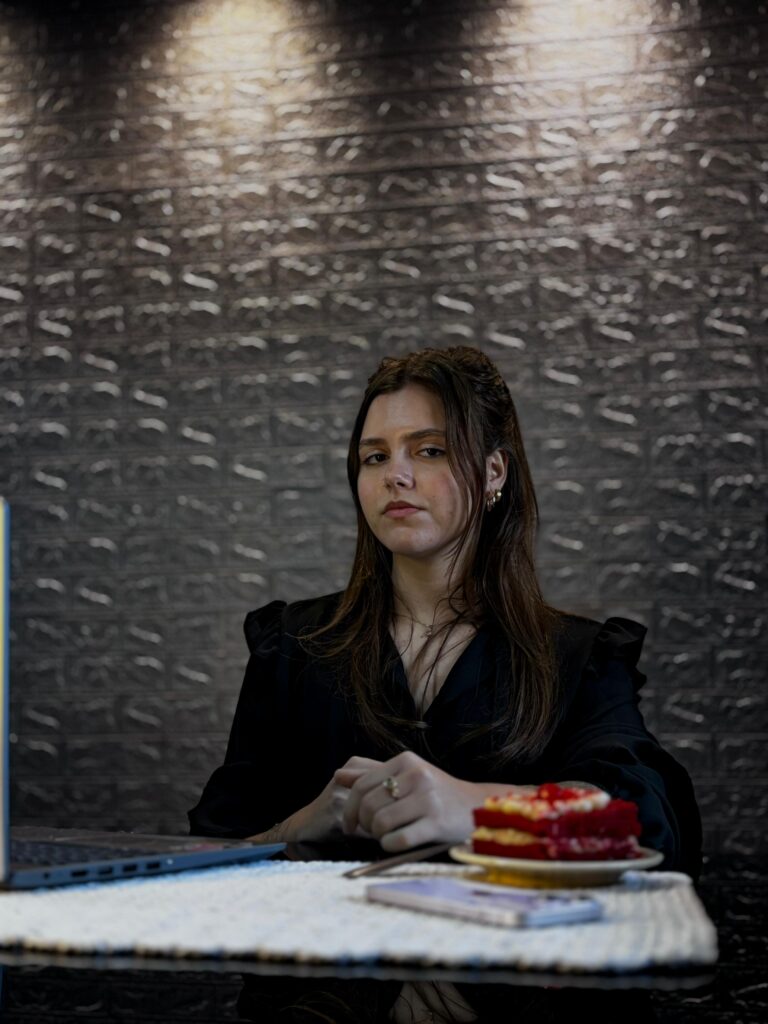 Kamila Lais Kuhn sentada com notebook e bolo rosa ao lado, expressão séria, usando roupa formal, transmitindo autoridade e expertise em nutrição e emagrecimento.