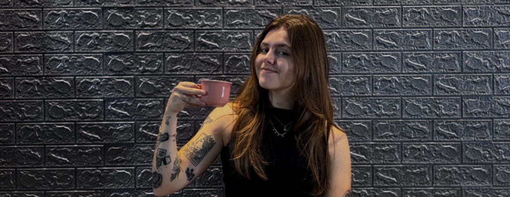 Kamila Lais Kuhn sorrindo levemente, segurando uma xícara de café ao lado do rosto, transmitindo equilíbrio, bem-estar e prazer na rotina.