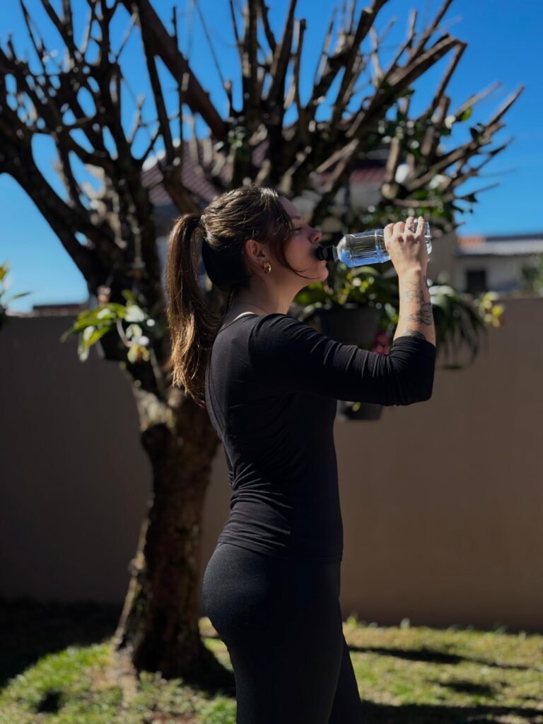 Kamila Lais Kuhn usando roupa de exercício e tomando água, mostrando seu estilo de vida ativo e envolvimento com esportes e bem-estar.