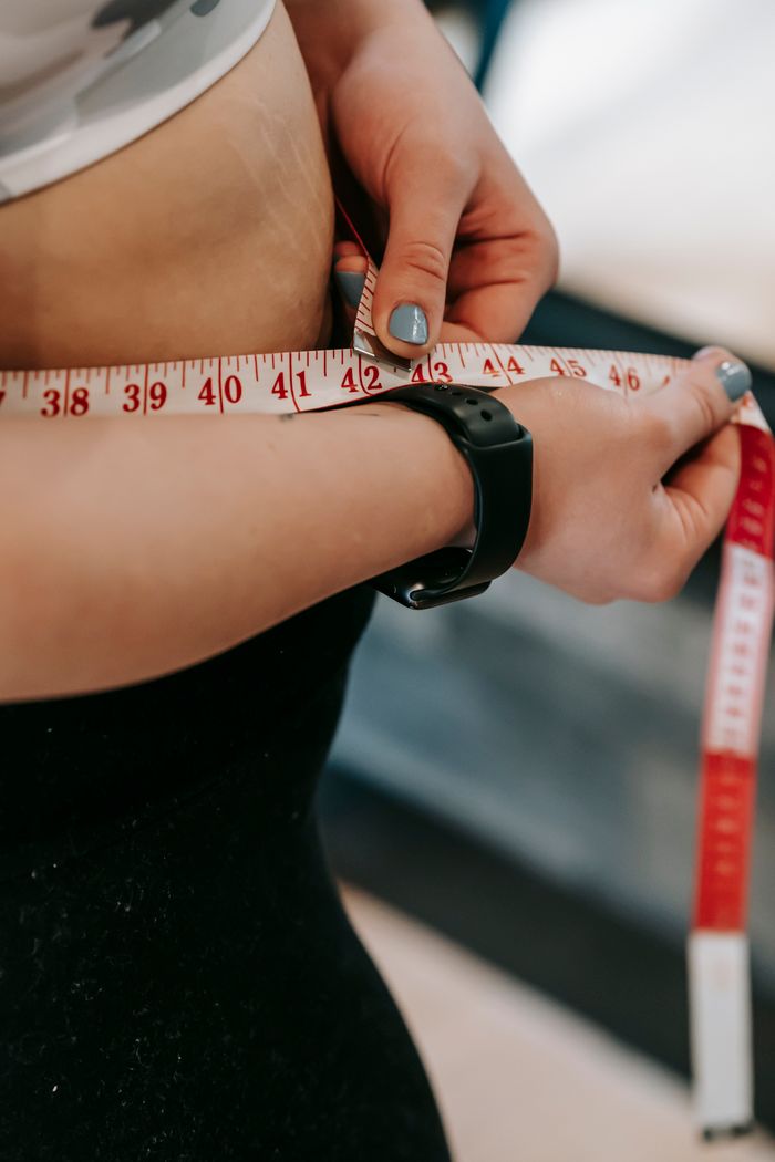Crop anonymous female with measuring tape around big belly on blurred background of gym